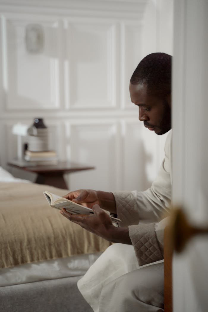 A man in a bathrobe sits in a cozy bedroom, deeply engaged in reading a book.